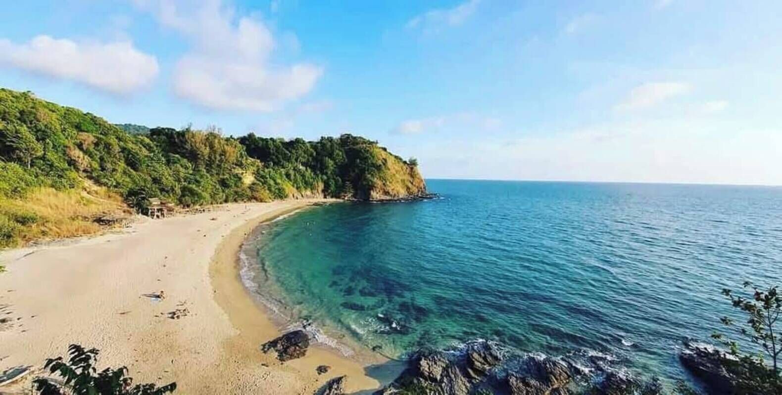 A coastal beach with clear blue waters, sandy shore, and a forested cliff.