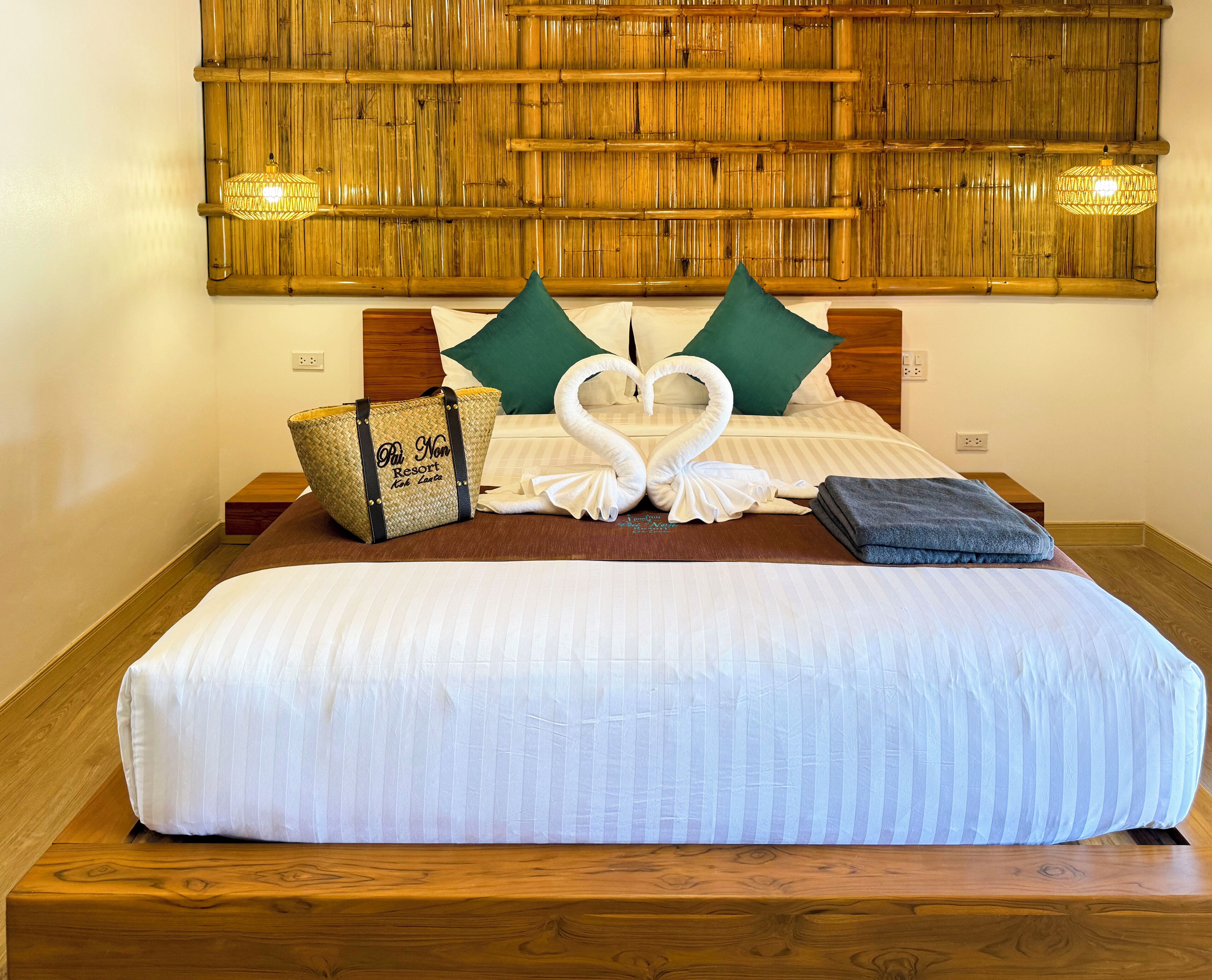 A neatly made bed with a heart-shaped towel arrangement, a woven bag, and a folded towel on the side.