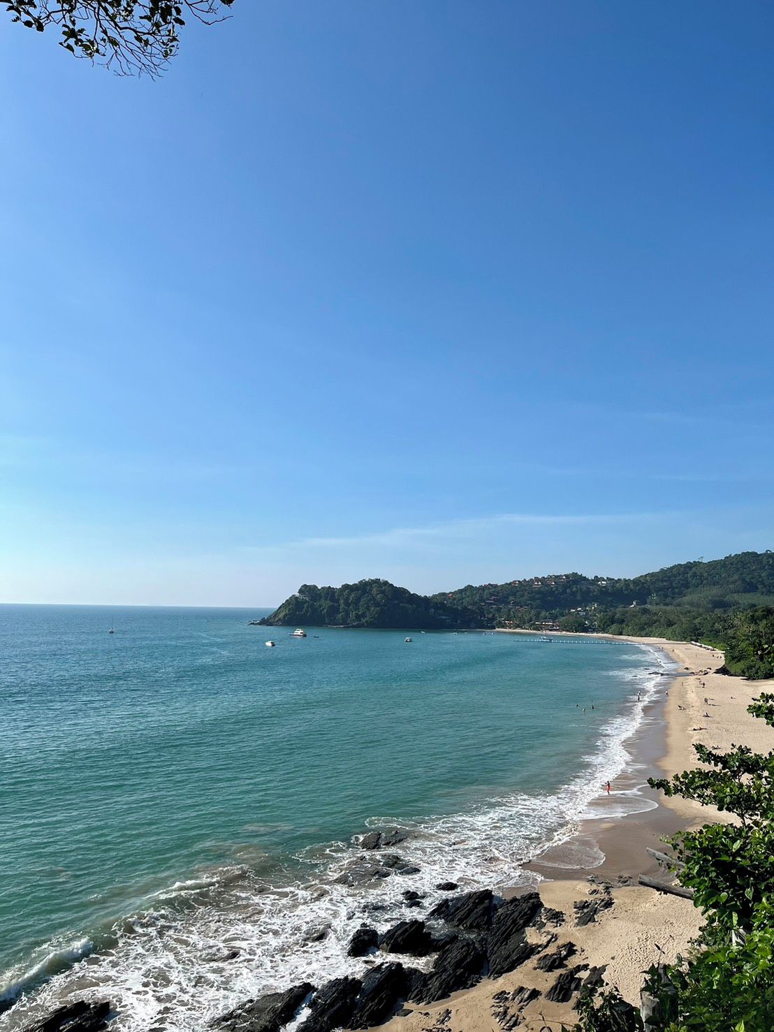 A coastal landscape with a sandy beach, rocky shoreline, and a distant hill with vegetation.