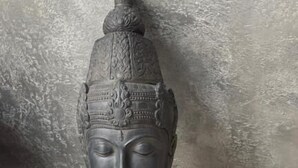 Une statue de Bouddha en pierre, ornée de sculptures complexes, est posée sur un plancher en bois, adossée à un mur à la texture particulière.