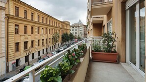 Un balcon orné d’une rangée de plantes en pot, d’où l’on peut admirer une rue bordée de voitures garées et d’immeubles.