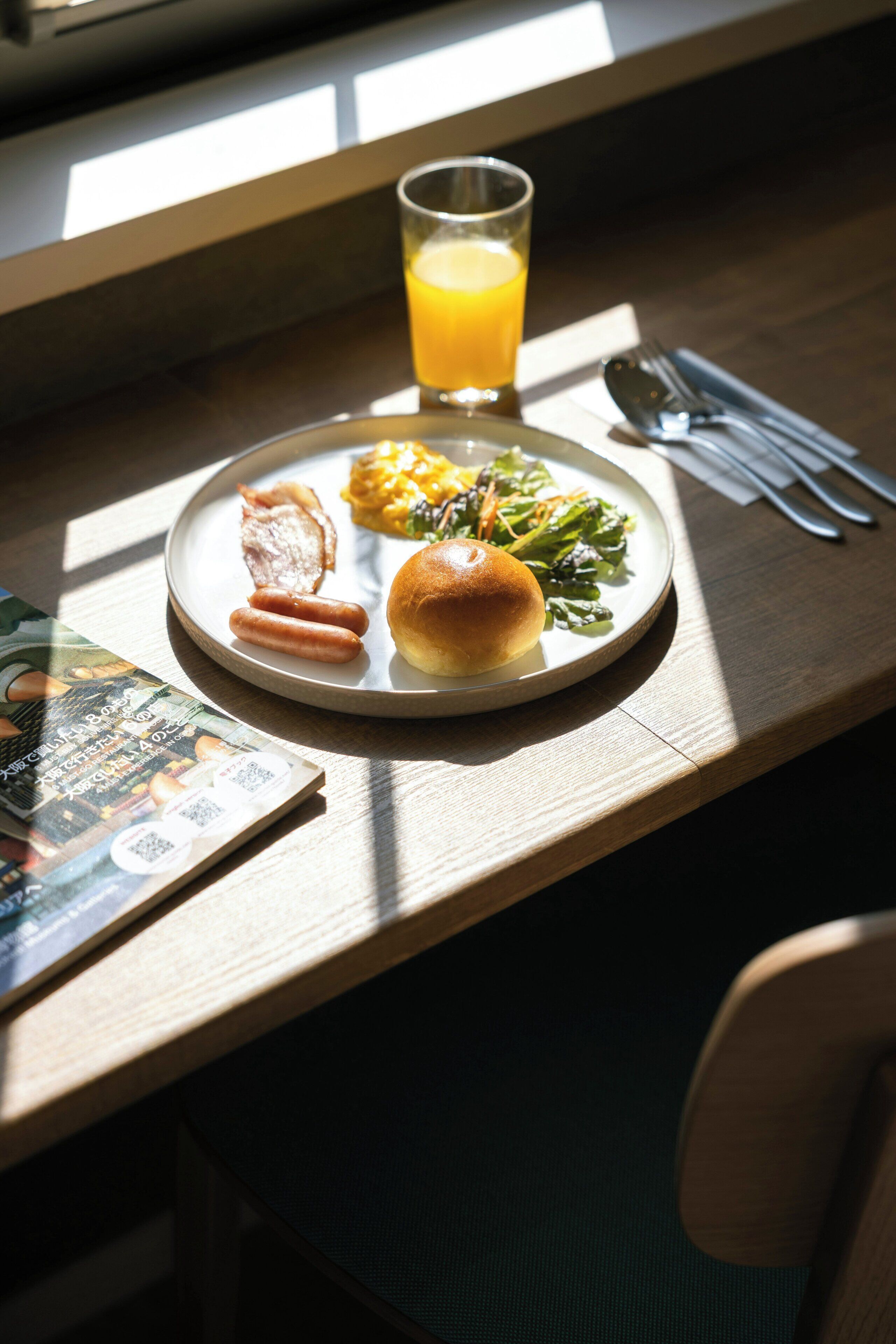 Une assiette de petit-déjeuner comprenant du bacon, des saucisses, un pain et des légumes verts, accompagnée d’un verre de jus d’orange, sur une table en bois avec une chaise et un menu.