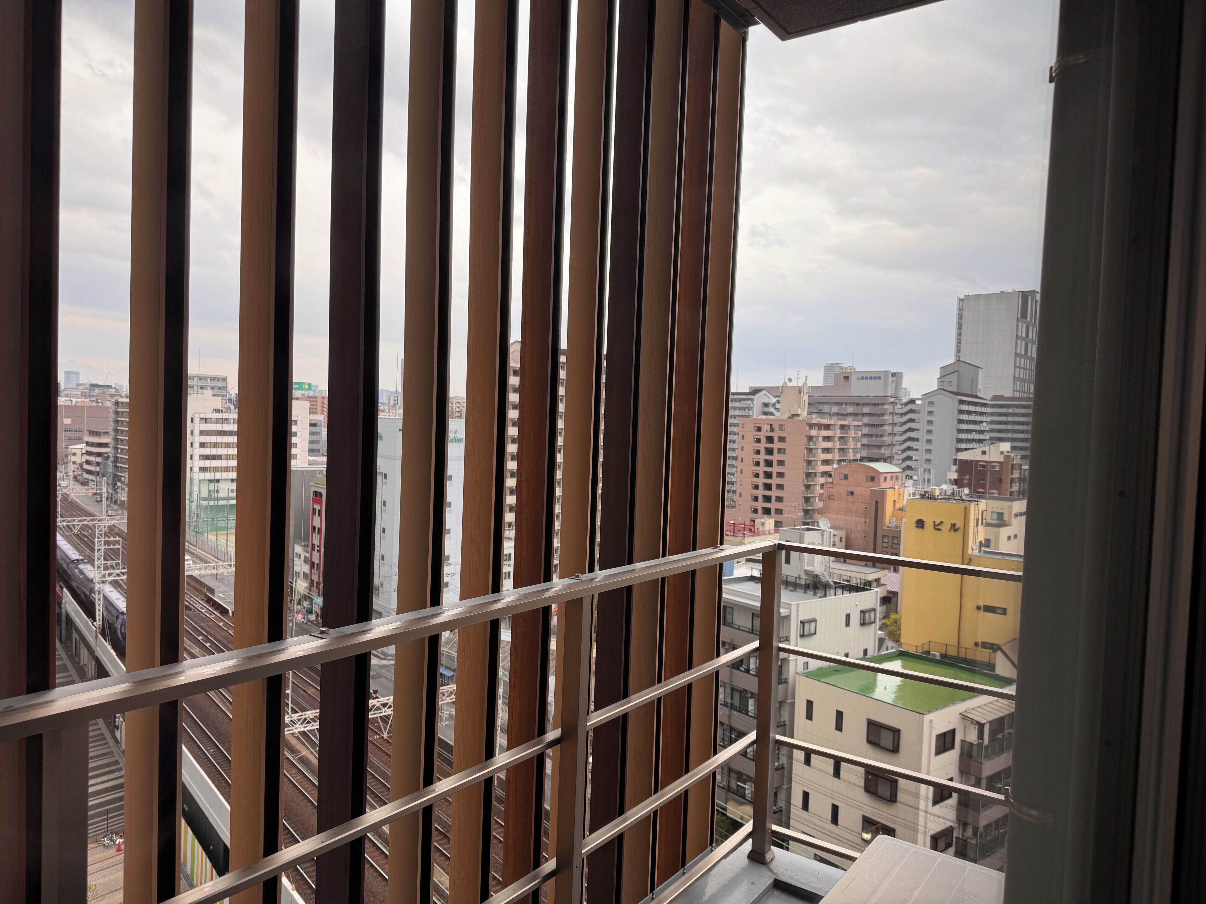 A view from a balcony with vertical wooden slats, overlooking a cityscape with buildings and a railway track.