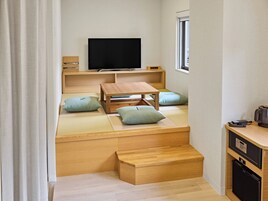 A traditional Japanese room with tatami flooring, a low wooden table, cushions, a television, and a small refrigerator.