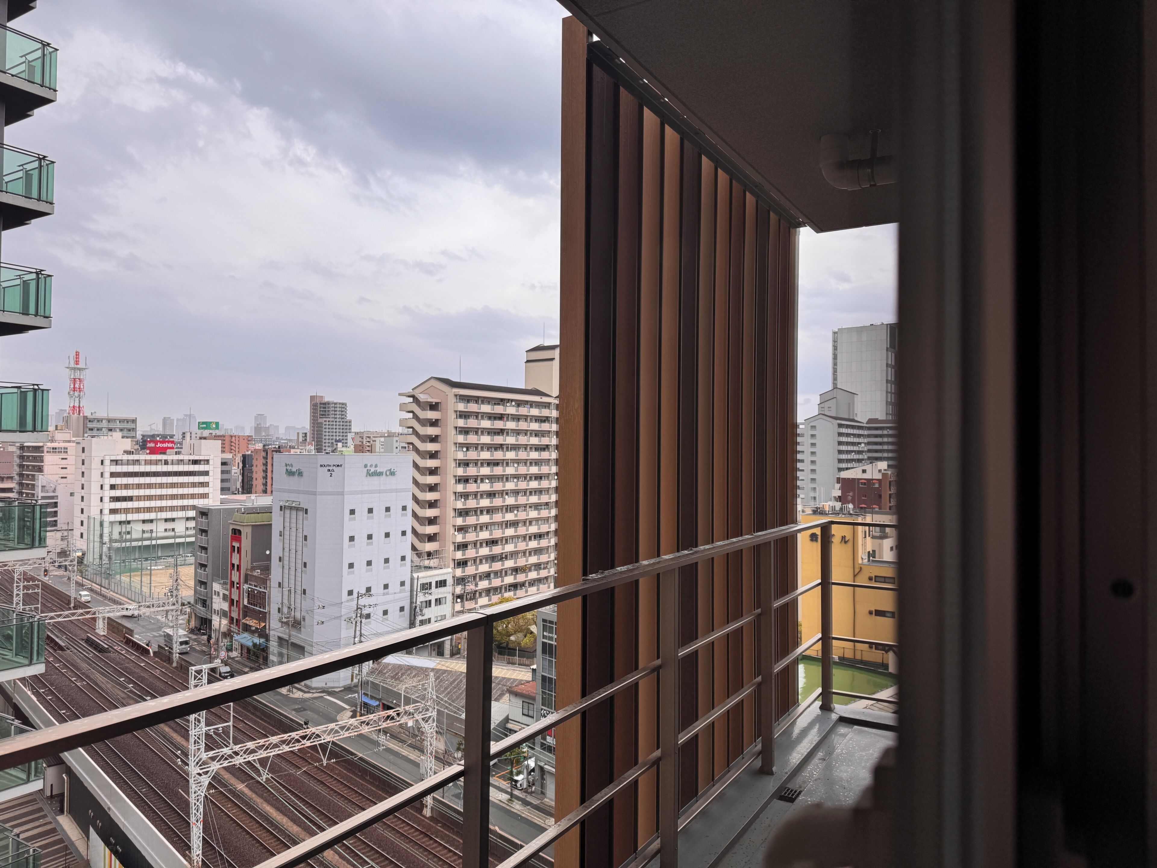 Blick auf eine Stadtlandschaft von einem Balkon mit Metallgeländer, mit Gebäuden und einer Bahnstrecke.