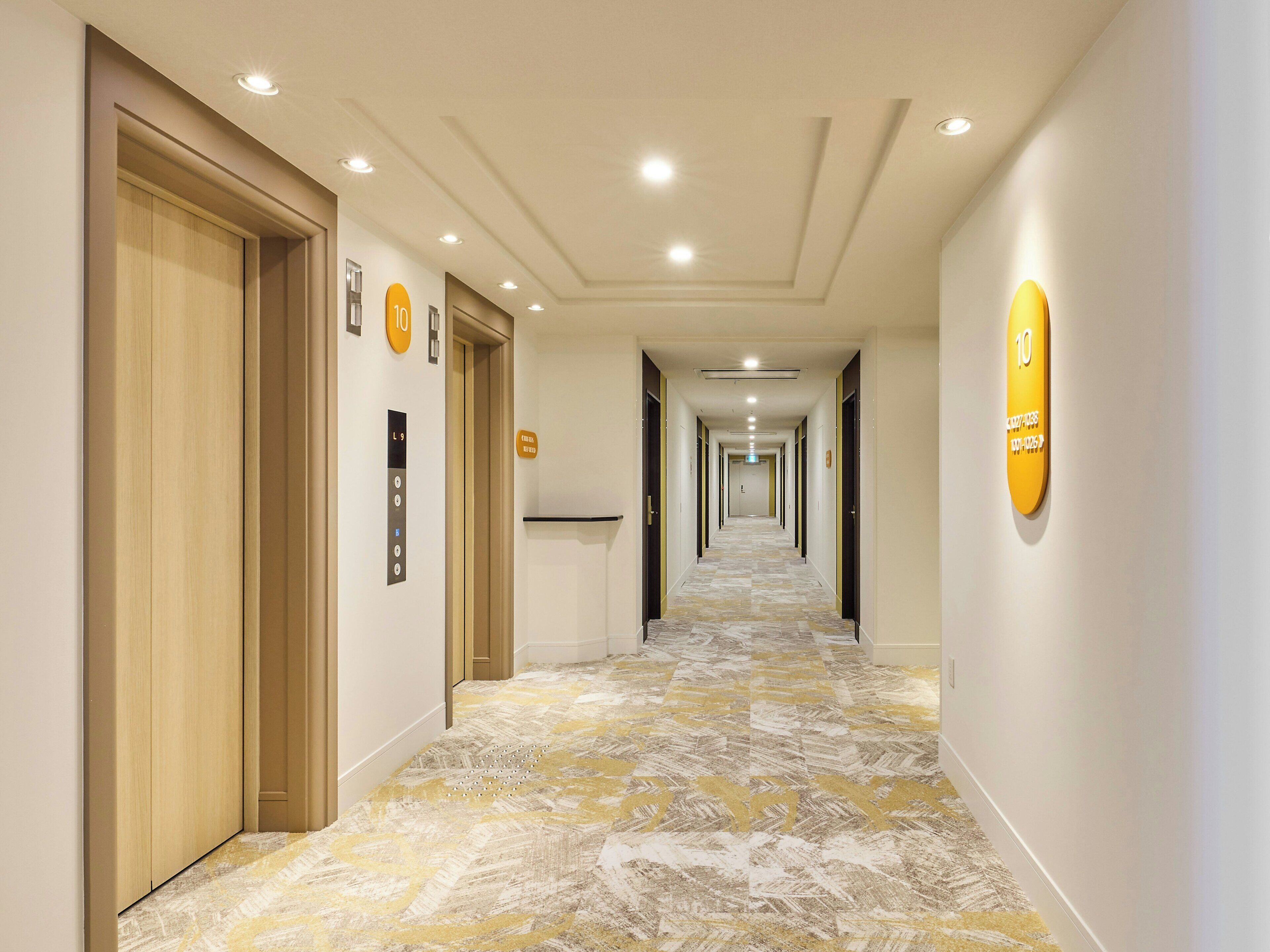 Un pasillo de hotel con puertas de madera, botones numerados del ascensor y una alfombra estampada.