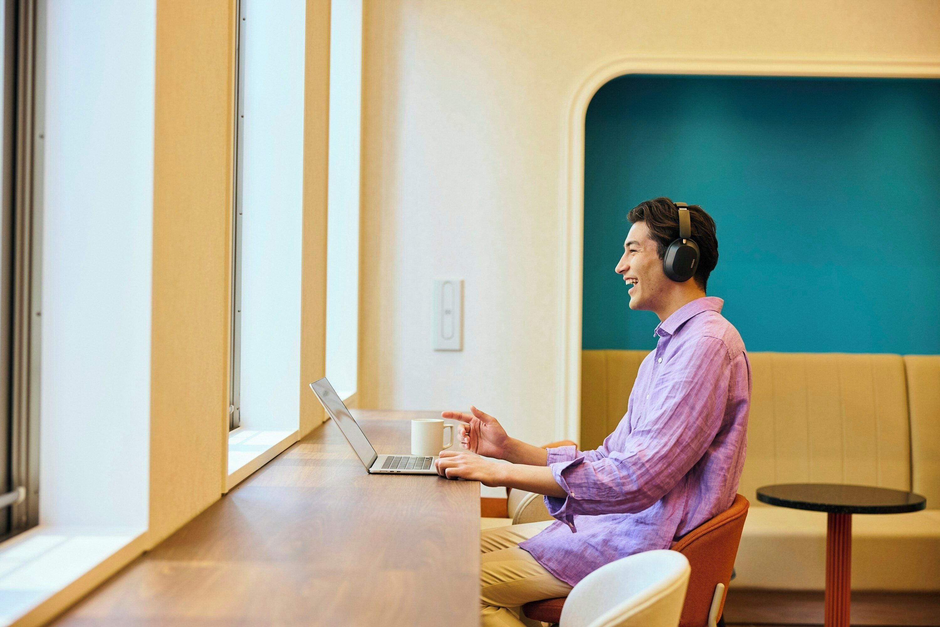 Una persona sentada a una mesa con una computadora portátil y una taza, en una habitación con ventana y pared color turquesa.