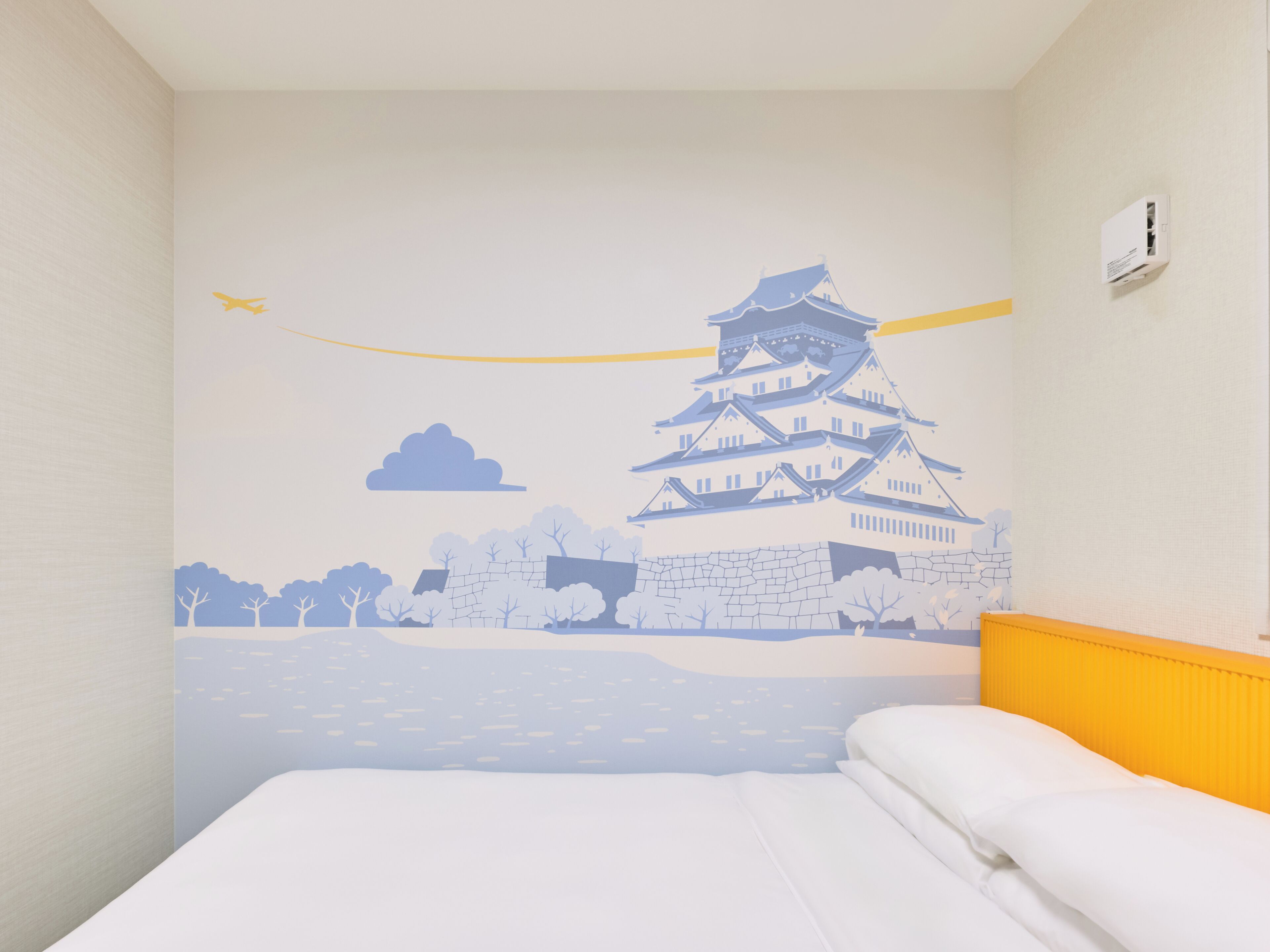A hotel room with a bed, a wall mural of a traditional Japanese building, and a small air conditioning unit on the wall.