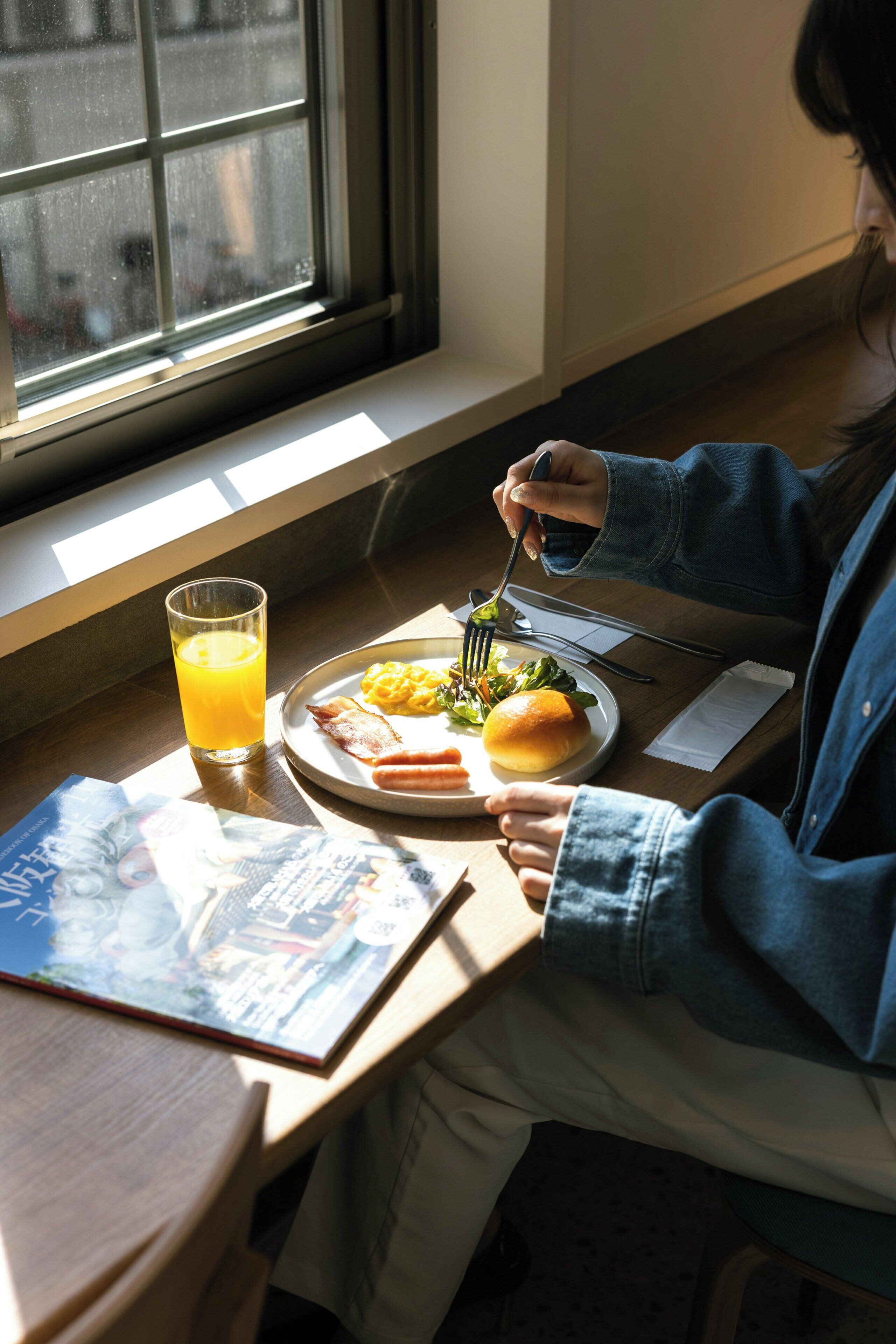 Iemand zit aan een tafel met een maaltijd, een glas sinaasappelsap en een tijdschrift.