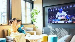 Ein Wohnzimmer mit Fernseher, der ein Sportspiel zeigt, einer Pflanze und einer Sitzecke mit Kissen.