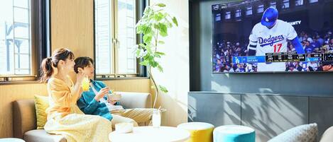 A living room with a TV displaying a sports game, a plant, and a seating area with cushions.