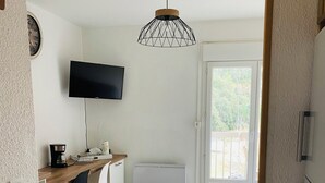 Une pièce de taille modeste, avec un bureau en bois, une chaise noire, un téléviseur fixé au mur, un luminaire au plafond et une porte-fenêtre coulissante donnant sur un espace extérieur.