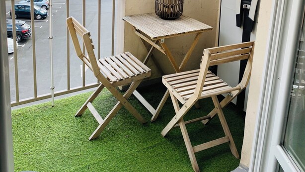 Un balcon avec une petite table et deux chaises, un pot de fleurs et une vue sur un parking et des collines lointaines.