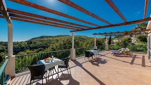 Um terraço com mesa posta para dois, uma cadeira e um espaço de estar com vista para uma paisagem montanhosa.