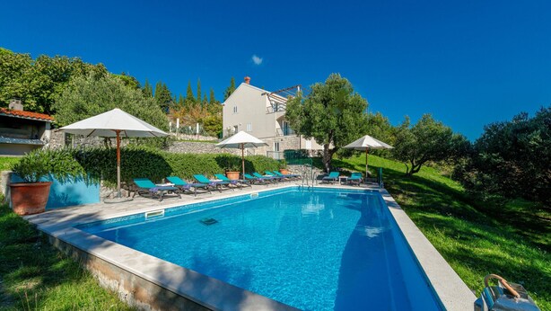 Uma piscina com espreguiçadeiras e guarda-sóis, cercada por vegetação e um edifício.