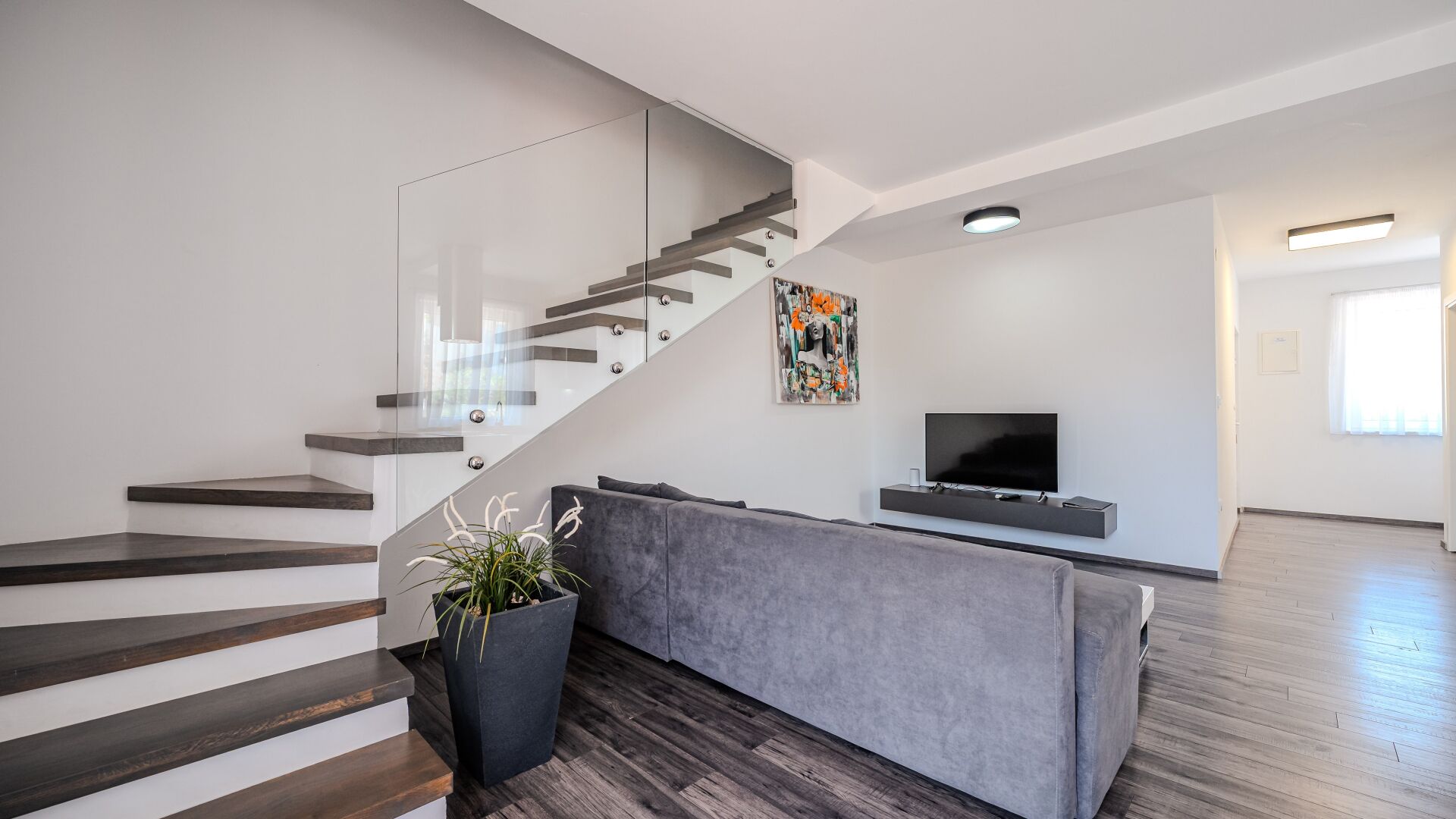 Ein modernes Wohnzimmer mit Treppe, einem grauen Sofa, einem Flachbildfernseher und einem Blumentopf.