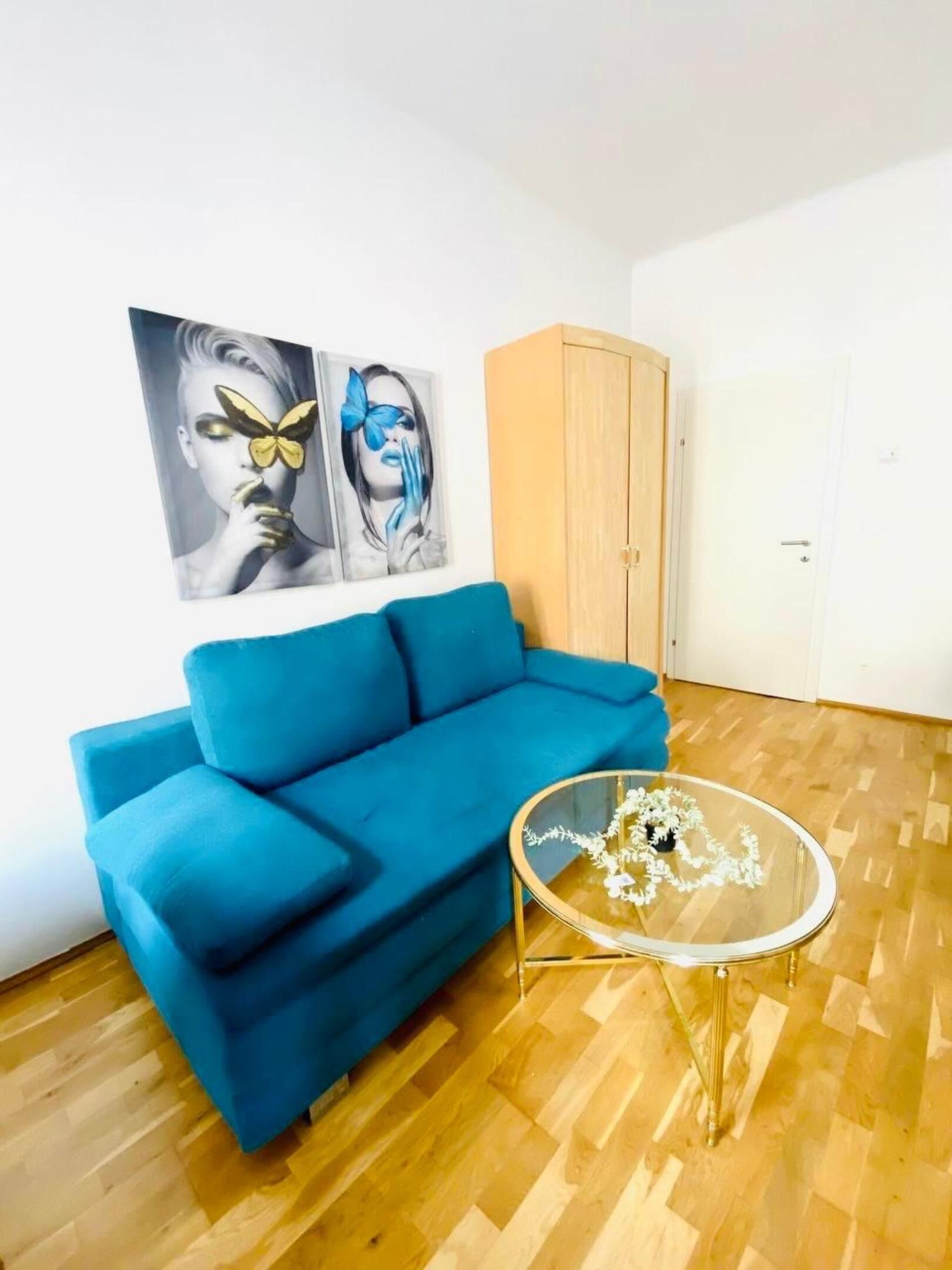 A room with a blue sofa, a glass coffee table with flowers, and two wall art pieces.