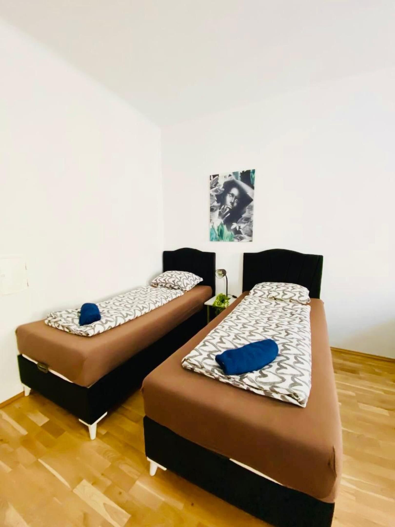 A room with two single beds, each with a brown mattress and patterned pillows. A small table with a lamp is between the beds.