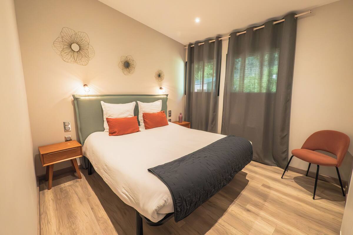 Ein ordentlich eingerichtetes Schlafzimmer mit einem großen Bett, einem Nachttisch, einem Sessel und einem Fenster mit Vorhängen.