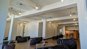 A spacious, modern interior with high ceilings and pendant lighting. There are multiple seating areas with bean bags and tables, and a visible kitchen area in the background. - Oak Ray La Ella Breeze (Ella)