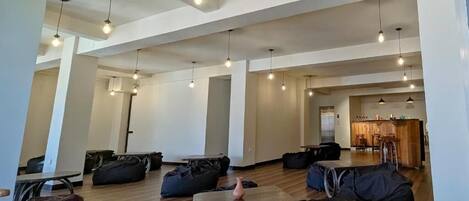 A spacious, modern interior with high ceilings and pendant lighting. There are multiple seating areas with bean bags and tables, and a visible kitchen area in the background.