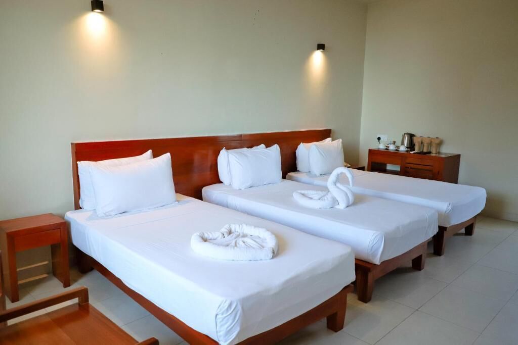 A hotel room with two single beds, each with a wooden headboard, white linens, and towels arranged on the beds. There is a wooden nightstand with a lamp and a small table with a kettle and cups.