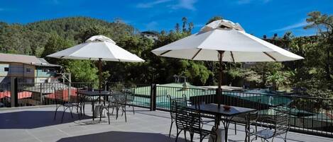 An outdoor patio with tables and chairs, large umbrellas, and a pool in the background.
