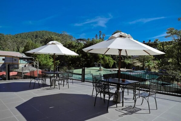 Un patio al aire libre con mesas y sillas, grandes sombrillas y una piscina al fondo.