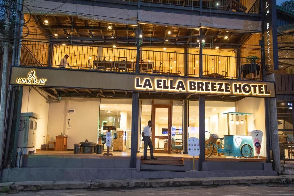 The exterior of La Ella Breeze Hotel with a visible sign, a balcony with seating, and a bicycle parked outside.