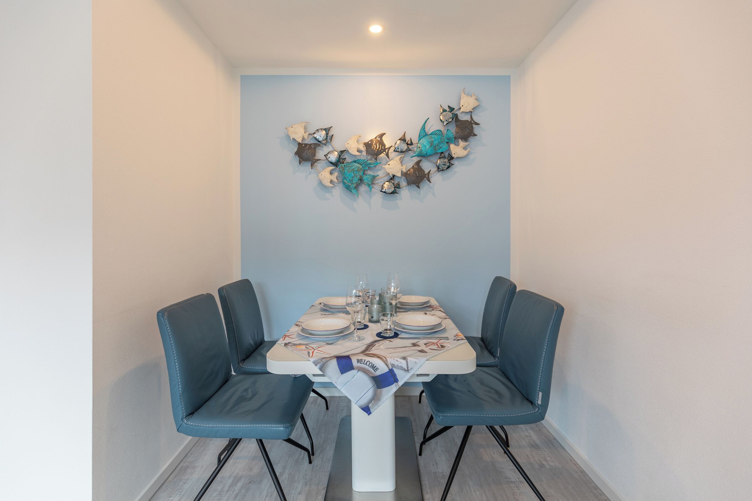 Un comedor con una mesa dispuesta para cuatro personas, sillas tapizadas en azul y una decoración de peces montada en la pared.