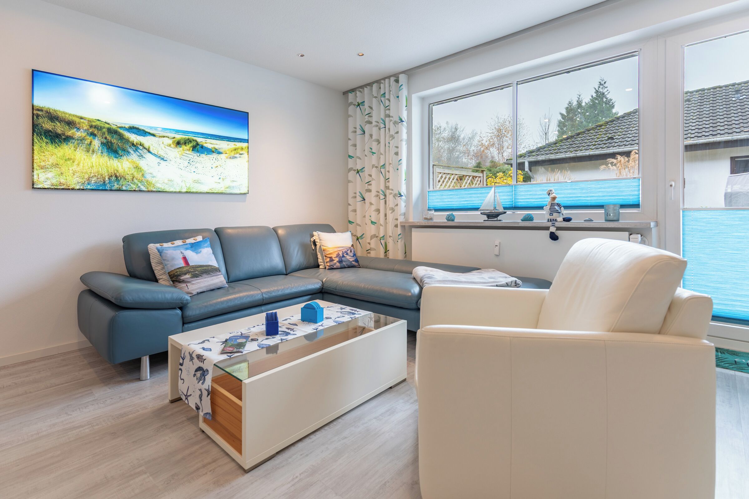 Een moderne woonkamer met een hoekbank, een witte fauteuil, een glazen salontafel en een groot raam met uitzicht op een blauw huis en groen.