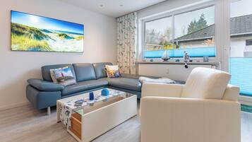 Een moderne woonkamer met een hoekbank, een witte fauteuil, een glazen salontafel en een groot raam met uitzicht op een blauw huis en groen.