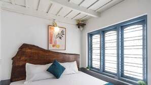 Ein Bett mit Holz-Kopfstück, weißer Bettwäsche und einer türkisnen Tagesdecke. Ein Fenster mit blauen Rollläden und eine Pflanze im Fensterbrett.