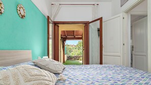 A bedroom with a patterned bedspread, a headboard, and a view through an open door to a garden. - Holiday Home 'Villetta Via Tempio' with Private Terrace, Private Garden and Air Conditioning (San Teodoro)