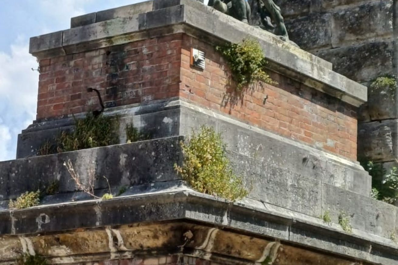 A stone monument with a statue on top, featuring a brick base and some vegetation growing around it.