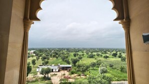A view through an arched window with a view of a rural landscape, including a building and greenery. - Abhushan Mahal (Degana)