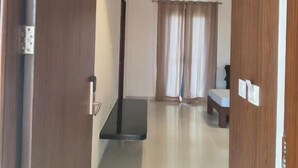 A hotel room with a wooden door, a small table, a bench, and a window with curtains. - Abhushan Mahal (Degana)