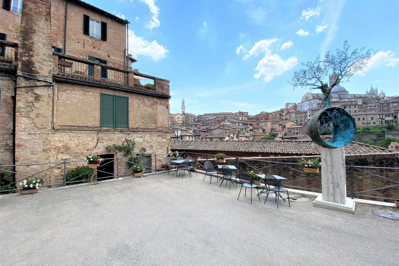 Eine Terrassengruppe mit Tischen und Stühlen, eine dekorative Baumskulptur und im Hintergrund eine Ansicht historischer Gebäude.