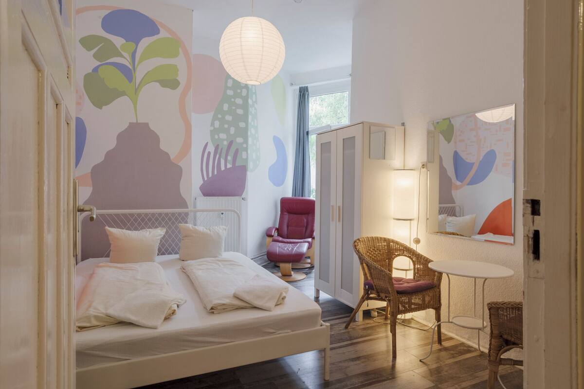 A bedroom with a bed, a red chair, a wicker chair, a small table, and abstract wall art.