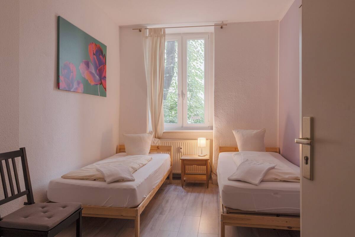 A small, well-lit room with two single beds, a wooden chair, a radiator, and a window with curtains.