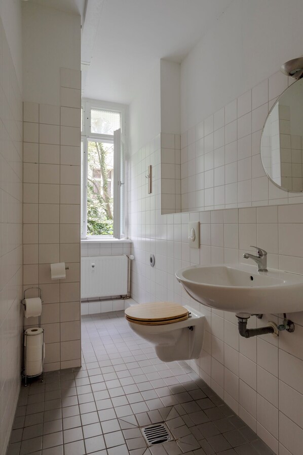 A small, clean bathroom with white tiles, a wooden toilet, a white sink, and a window providing natural light.