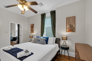 A hotel room with a bed, a ceiling fan, a window with curtains, a desk with a lamp, and artwork on the walls. - Classic 2BR Mid-City shotgun with fenced yard. Walk to streetcar, near FQ & City Park. (New Orleans)