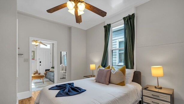 Ein Schlafzimmer mit Bett, Nachttischlampen, Deckenventilator, einem Fenster mit Vorhängen und Blick auf einen anderen Raum.