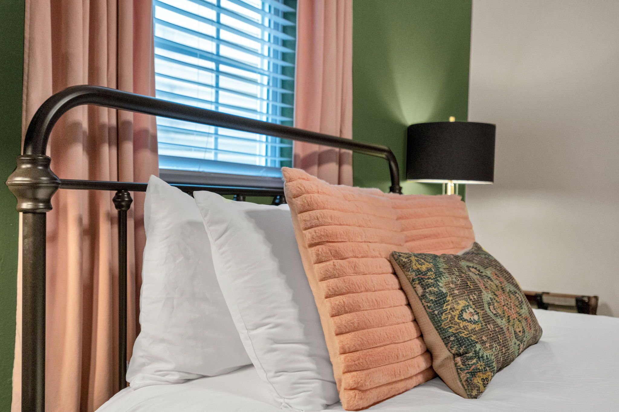 Un lit avec des oreillers blancs et roses, une lampe noire et une fenêtre avec des stores.
