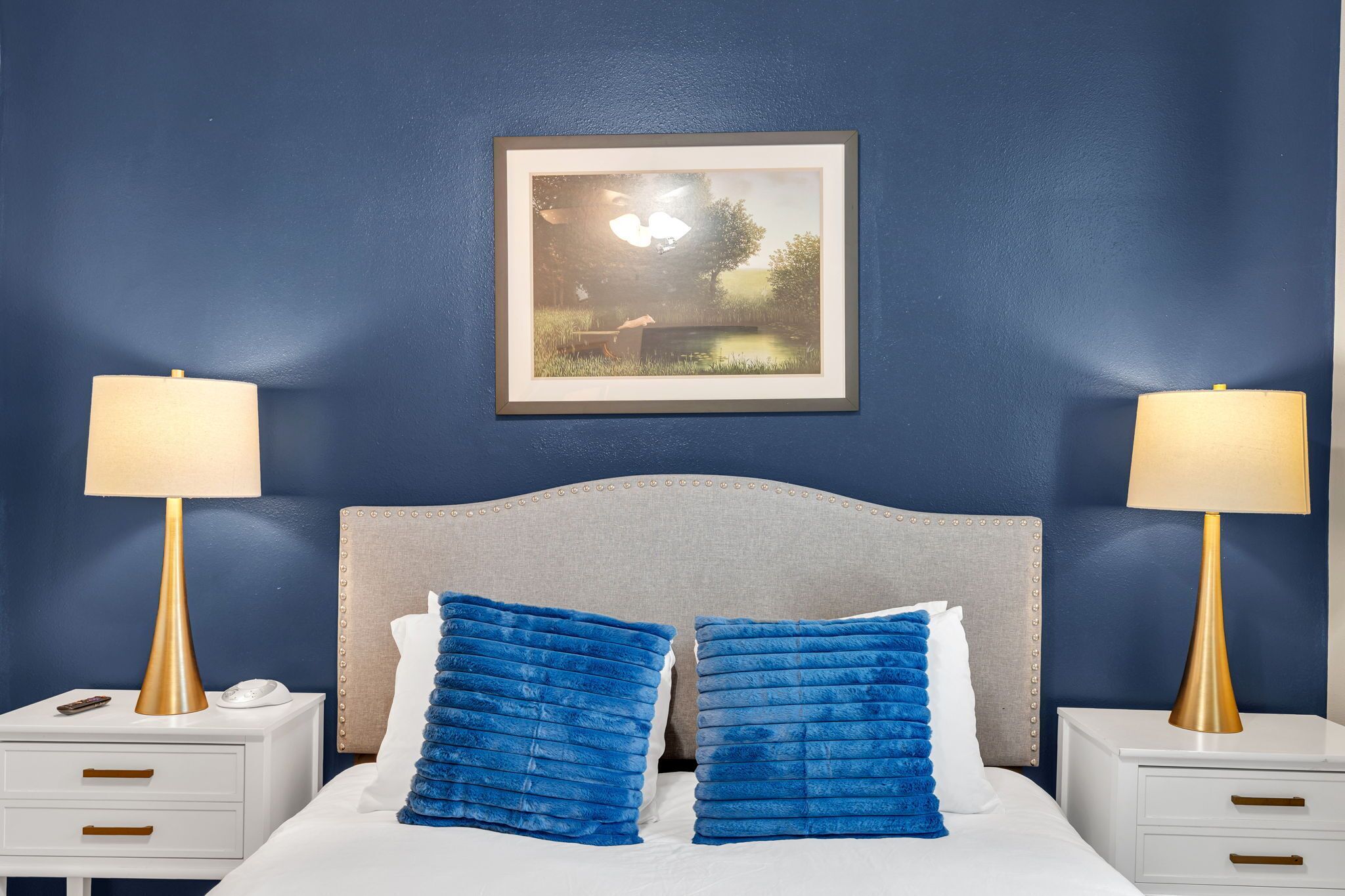 Une chambre avec un mur bleu, un tableau encadré, deux lampes de chevet, un lit avec des oreillers bleus et une table de chevet sur laquelle se trouvent un téléphone et une lampe.