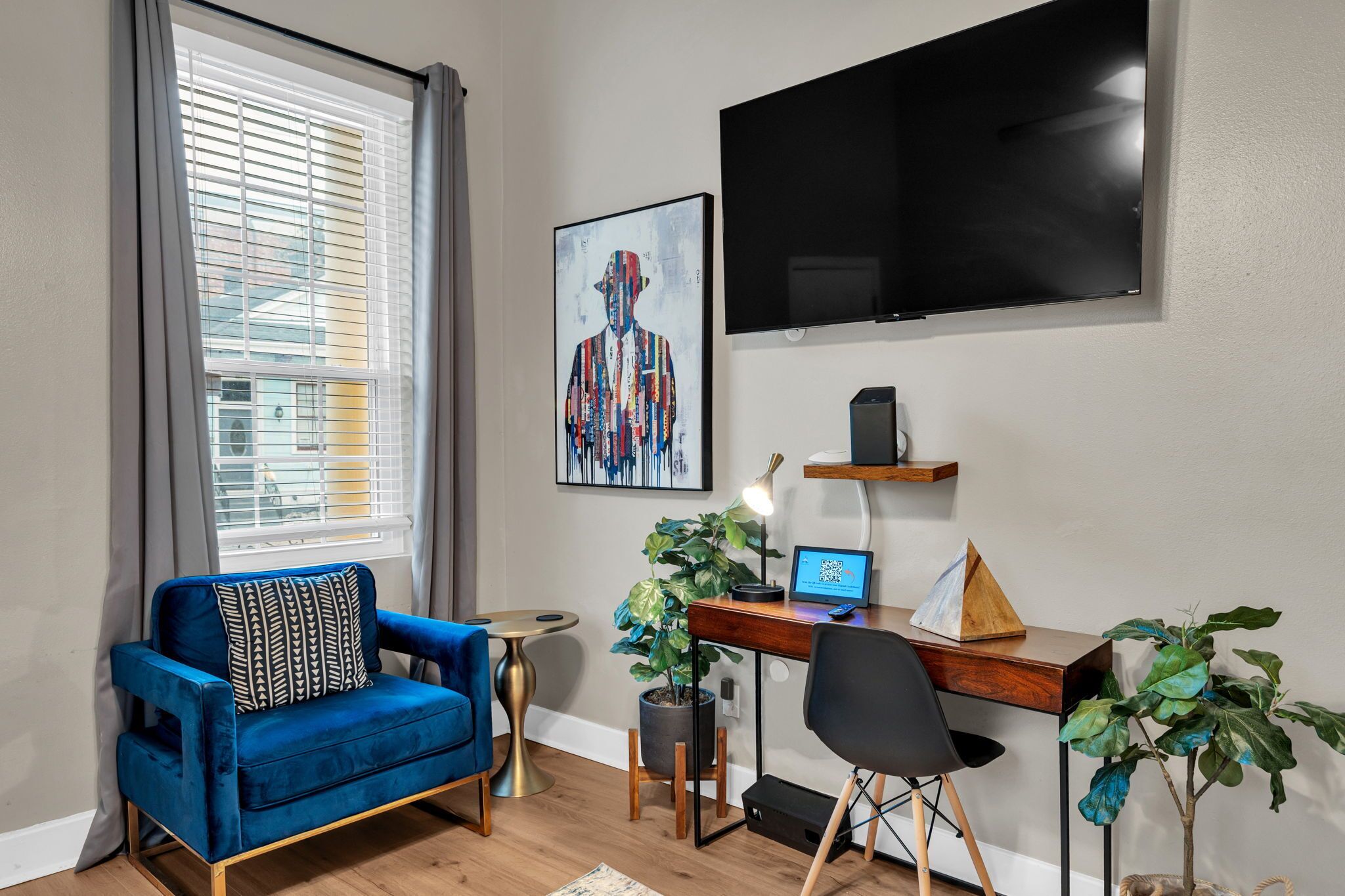 A modern living room with a blue sofa, a desk with a laptop, a TV, and a large window with blinds.