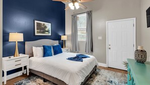 Une chambre à coucher comprenant un lit, des tables de chevet, un ventilateur de plafond, une fenêtre avec des rideaux, une porte et un vase décoratif posé sur une table.