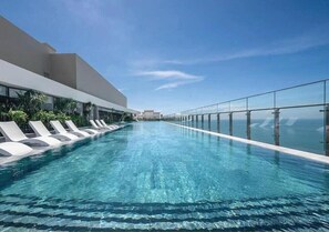 A modern infinity pool with clear blue water, surrounded by white lounge chairs and glass railings, overlooking a scenic view. - The Song Apartment - Havilla Vung Tau  (Ho Chi Minh City)