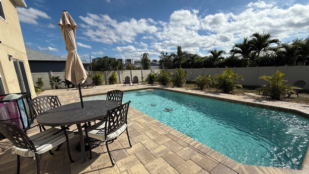 A swimming pool with a patio area, including a table and chairs, surrounded by palm trees and a fence.