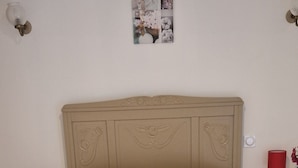 Un lit recouvert d’une couvre-lit à motifs floraux, une tête de lit ornée de sculptures, une petite table de chevet avec une lampe rouge et un tableau accroché au mur.